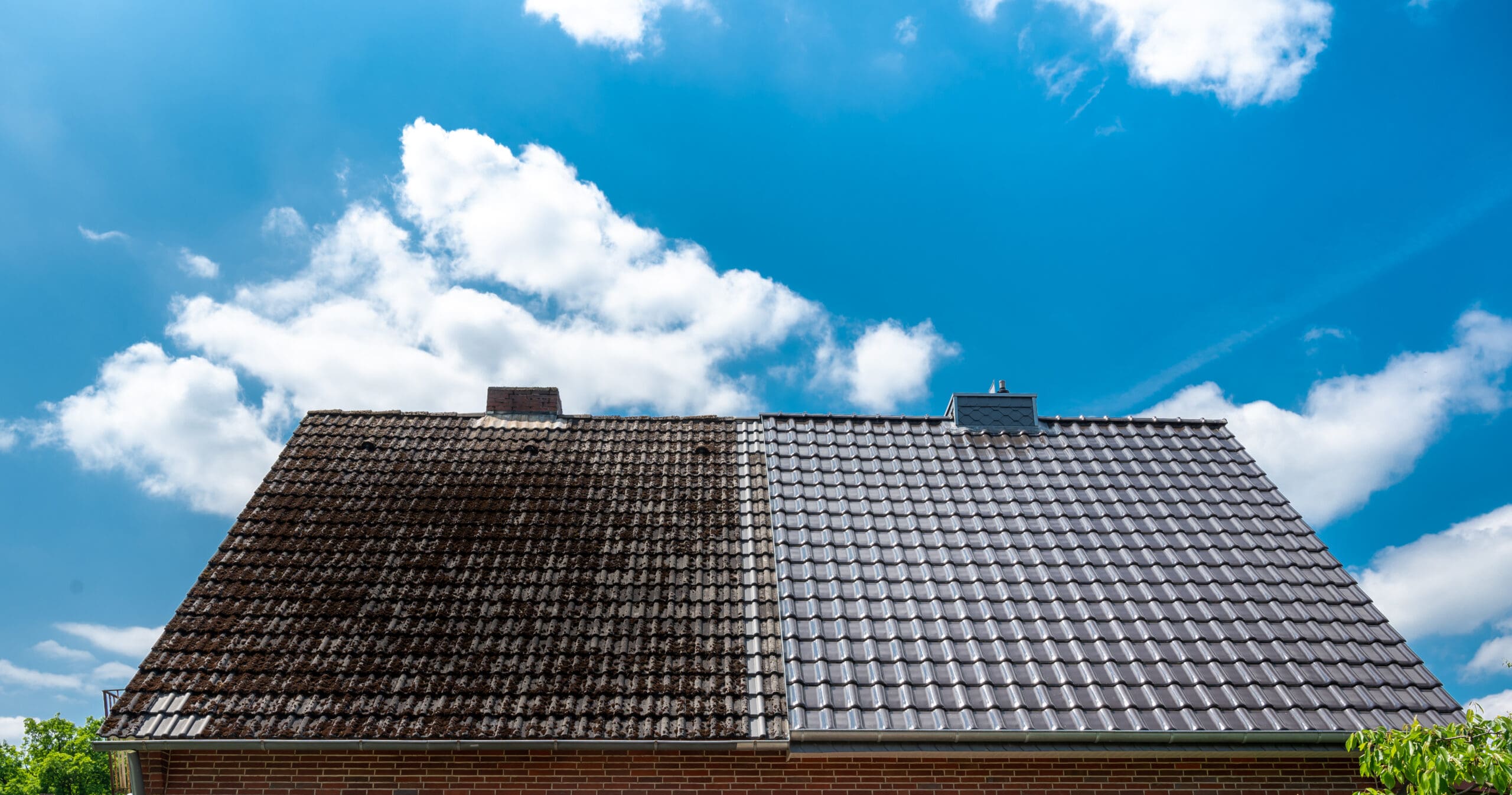 A house roof with two contrasting halves: the left side is covered in mold and dirt, appearing brown and weathered, while the right side is clean and bright with new shingles. A bright blue sky with scattered white clouds is in the background.