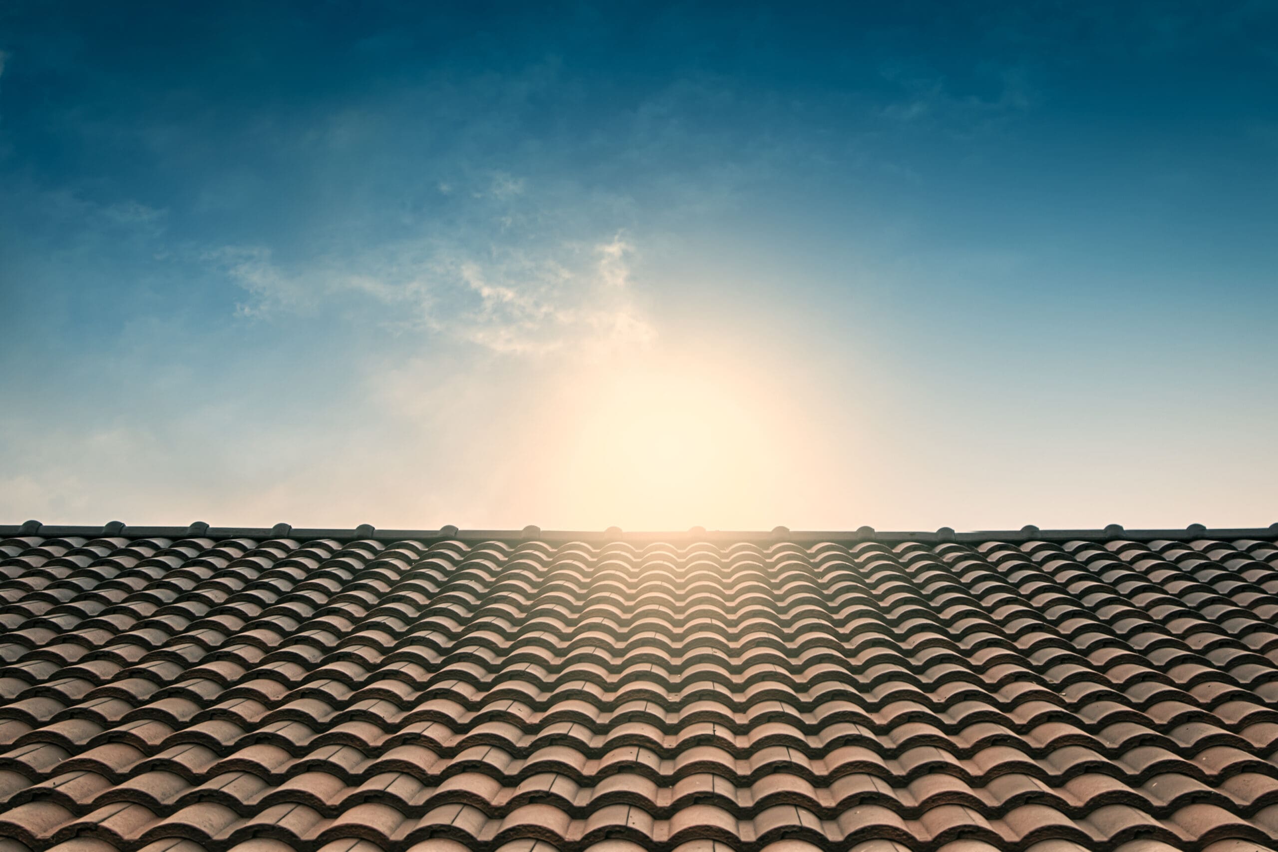 Sunrise over a tiled roof with a clear sky. The sun is partially hidden behind the roof, creating a bright glow and soft lens flare. The roof tiles form a uniform pattern, and the sky transitions from a deep blue to a light gradient near the horizon.