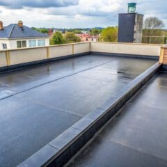 Flat rooftop with a smooth, black asphalt surface. Surrounding low walls frame the roof. In the background, a cloudy sky hovers over neighboring buildings and greenery. A small red brick chimney and a water tank with a hose are on the side.