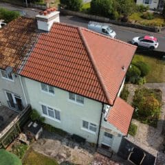 Aerial view of a semi-detached house with a red-tiled roof on the right and a weathered roof on the left. A white van and a red car are parked on the street. The yard features a small patio and garden area with green shrubs.
