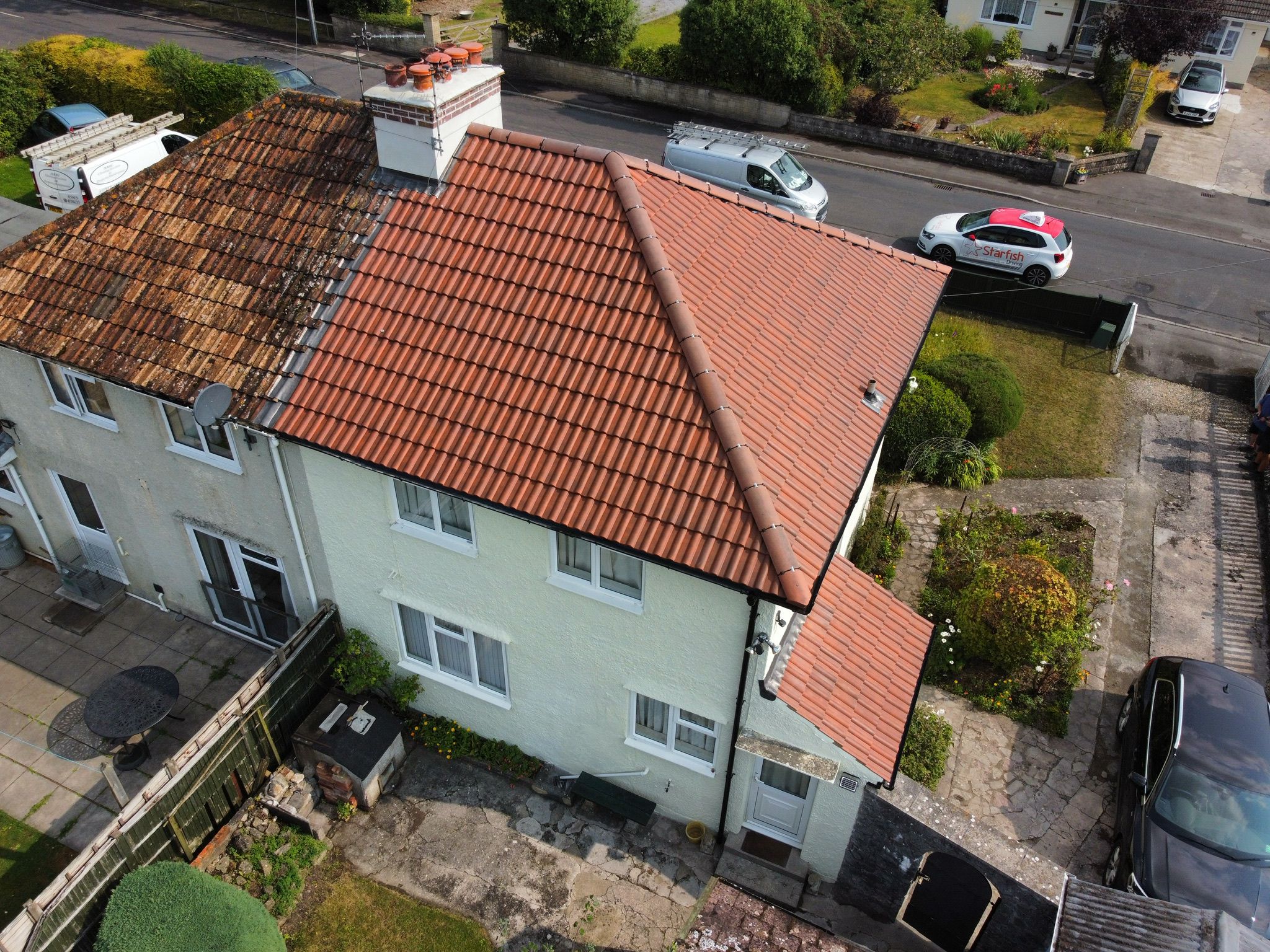Aerial view of a semi-detached house with a red-tiled roof on the right and a weathered roof on the left. A white van and a red car are parked on the street. The yard features a small patio and garden area with green shrubs.