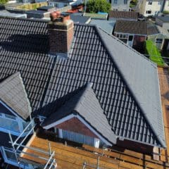 Aerial view of a house with new dark gray roof tiles and a red brick chimney. Scaffolding surrounds the building, indicating ongoing construction or renovation. Nearby rooftops and a green lawn are visible.