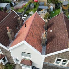 Aerial view of a house with a red-tiled roof, multiple chimneys, and an adjoining similar house. The backyard features a trampoline, lawn areas, and small outbuildings. Garden fences separate it from neighboring properties.