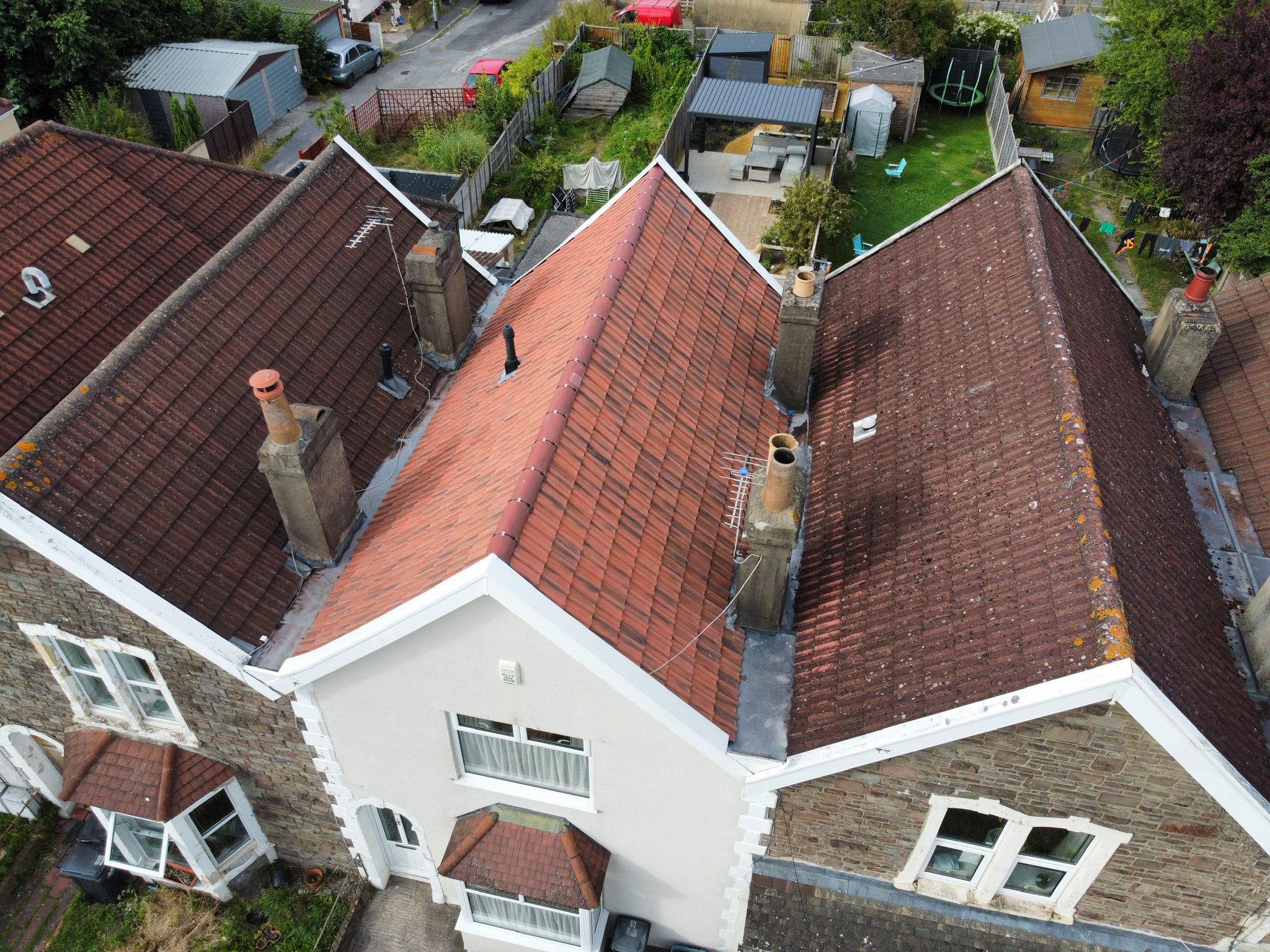 Aerial view of a house with a red-tiled roof, multiple chimneys, and an adjoining similar house. The backyard features a trampoline, lawn areas, and small outbuildings. Garden fences separate it from neighboring properties.