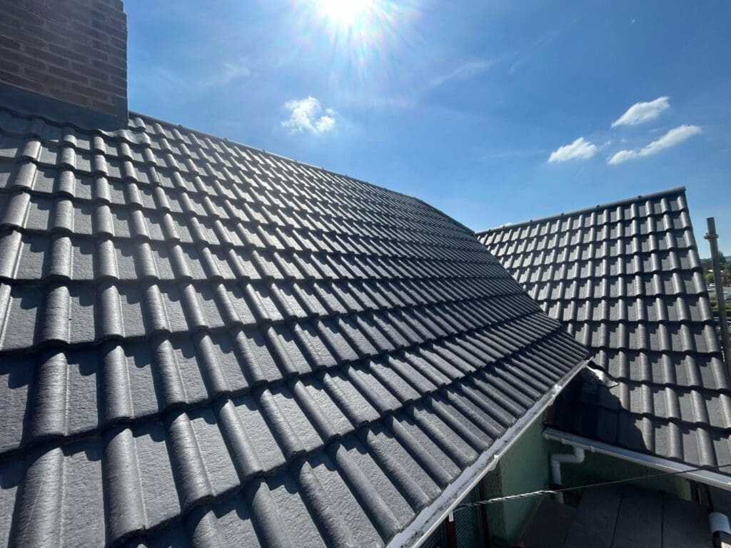 A sunny day view of a house roof with textured gray tiles, a chimney on the left, and clear blue sky above. The sunlight reflects off the roof, creating a warm and bright atmosphere.