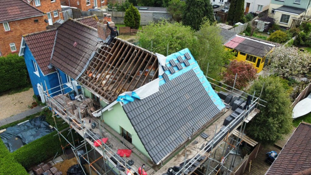 Aerial view of a house under construction. A worker on the roof is repairing or replacing tiles. Scaffolding surrounds the structure, and a blue tarp covers part of the roof. The area is residential, with trees and neighboring houses visible.