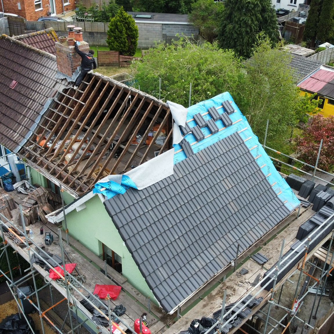 A house under renovation with a partially completed roof. One half is covered in new dark tiles, and the other half exposes wooden beams. A blue tarp covers part of the unfinished section. Scaffolding surrounds the house. Trees and buildings are in the background.