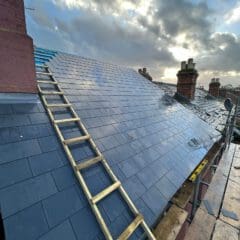 A newly installed slate roof on a house with a wooden ladder leaning against it. The sky is cloudy, and there are several chimney stacks visible in the background. A scaffolding platform is set up around the roof.