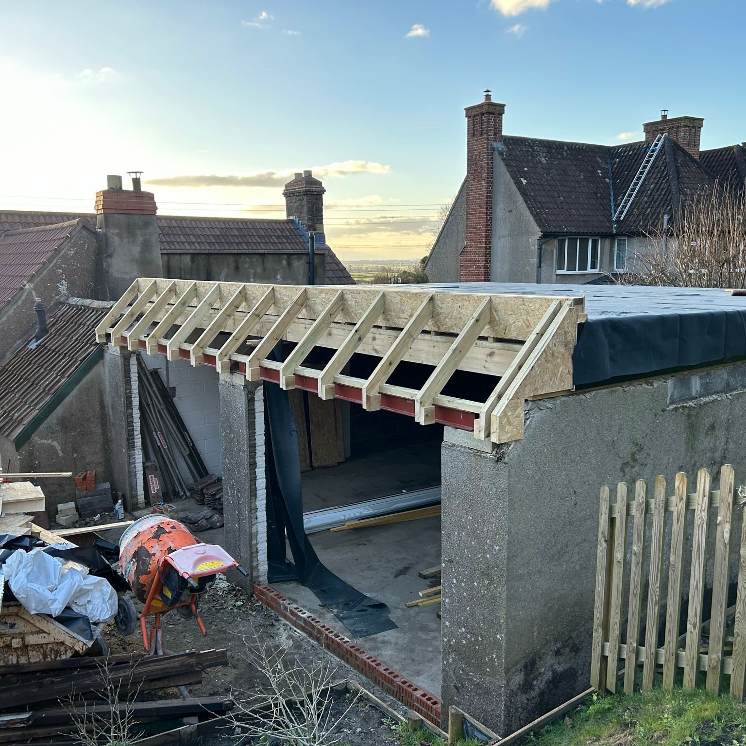 A partially constructed garage roof with exposed wooden beams and an unfinished structure. A wheelbarrow and building materials are scattered on the ground. Nearby houses with sloped roofs are visible under a partly cloudy sky.