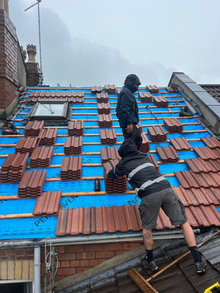 Two people wearing hooded jackets work on a sloped roof covered with blue tarpaulin and red tiles. One person stands, while the other kneels, adjusting tiles. A cloudy sky and chimney are visible in the background.
