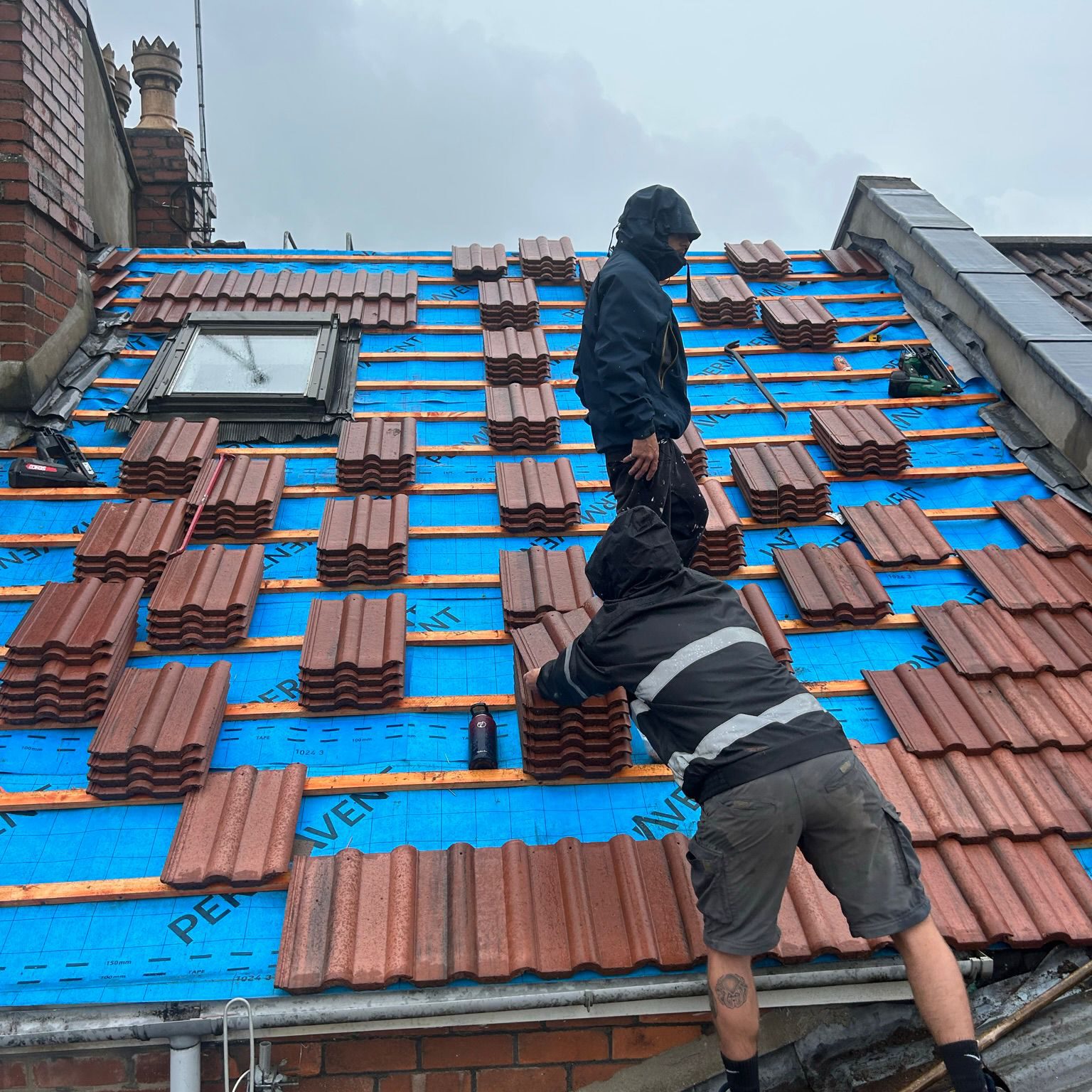 Two workers dressed in rain gear are positioning red roof tiles on a sloped roof with a blue underlayment. The overcast sky suggests rainy weather. A chimney is visible in the background.