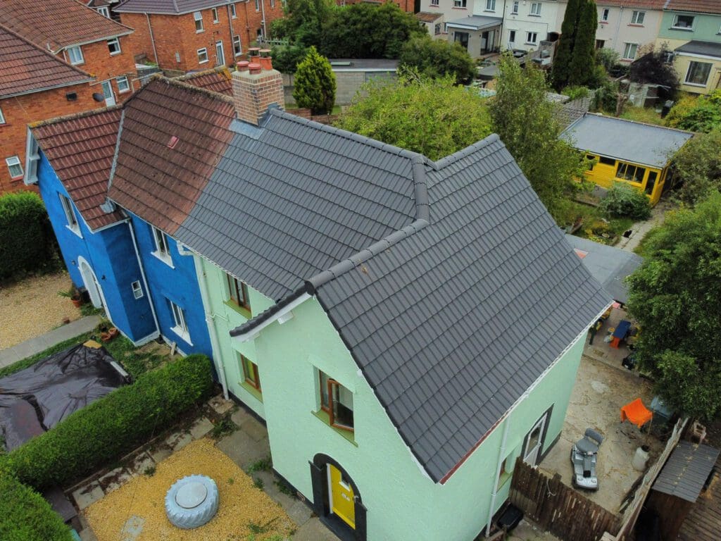 Aerial view of a semi-detached house with a newly renovated, dark gray gabled roof. The house on the left is painted blue and the one on the right is light green. Surrounding houses and greenery are visible in the background.
