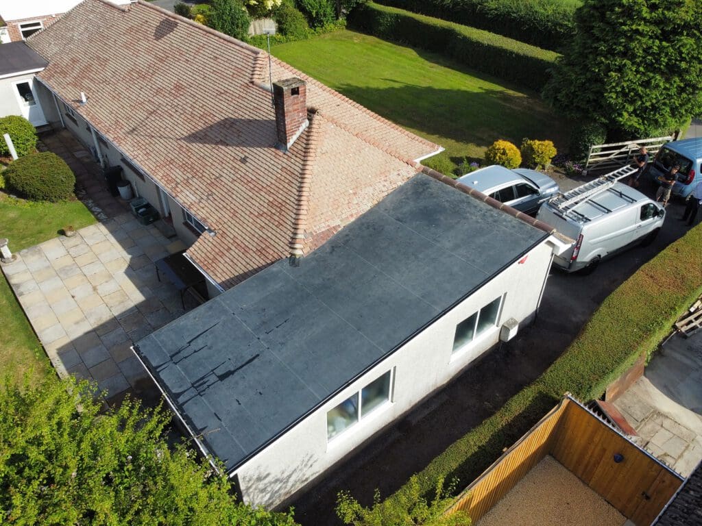 Aerial view of a house with a tiled roof and a flat-roofed extension. A driveway on the right has a white van and a person standing near it. The garden features neatly trimmed hedges and bushes. A patio area is visible on the left side.