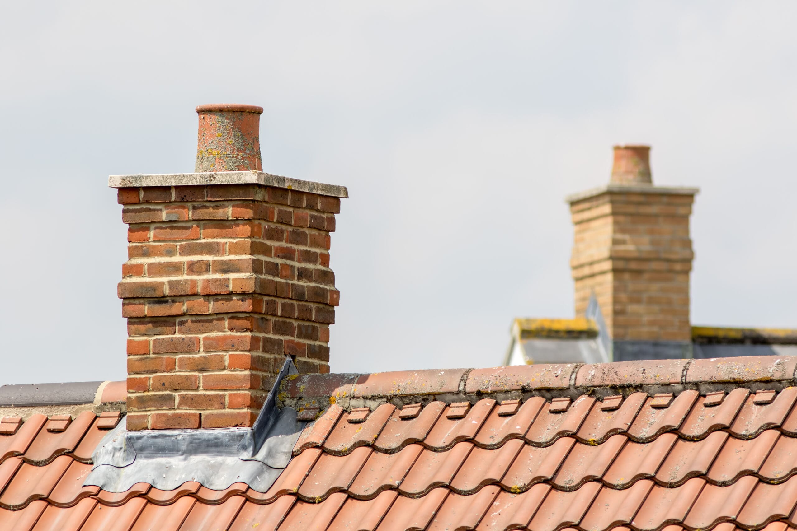 Two brick chimneys on a tiled roof, with moss-covered edges and a cloudy sky in the background. The roof is made of terracotta-colored tiles.