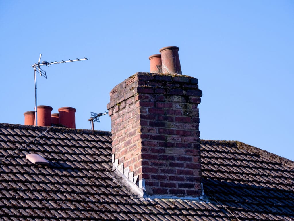 Brick chimney with two terracotta pots on a tiled roof. Two television antennas are also visible, one mounted near the chimney. The sky is clear and blue, providing a bright backdrop.