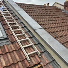 A rooftop with brown and reddish tiles, featuring a ladder leaning against it. The edge has gray metal flashing. Part of a person in blue clothing is visible near the ladder. The sky is overcast.