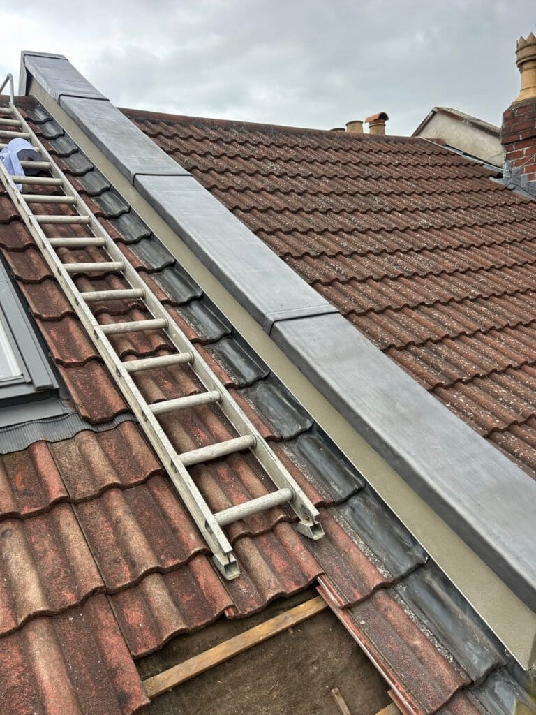 A rooftop with brown and reddish tiles, featuring a ladder leaning against it. The edge has gray metal flashing. Part of a person in blue clothing is visible near the ladder. The sky is overcast.