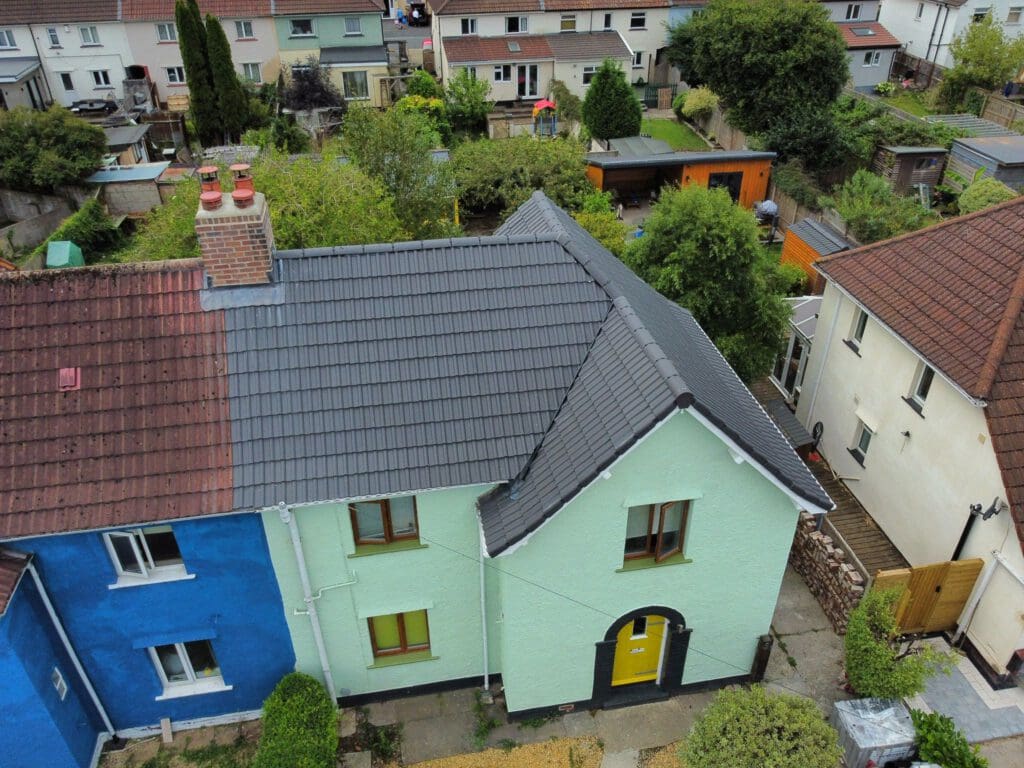 Aerial view of colorful houses. The left house has blue walls and a reddish-brown roof, the right house is light green with a dark gray roof and a yellow door. There are gardens with trees and shrubs in the backyards.