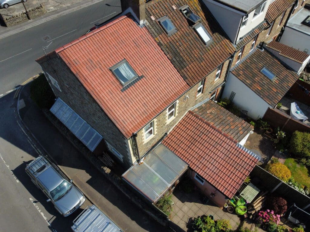 Aerial view of a two-story house with a red-tiled roof and stone exterior. There's a small garden with potted plants and a glass-roofed extension. A silver car is parked on the street corner with some refuse bins nearby.