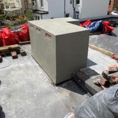 A rectangular, smooth-surfaced structure is on a rooftop surrounded by bricks, construction materials, and debris. Several red bags are stacked nearby, with residential houses visible in the background.