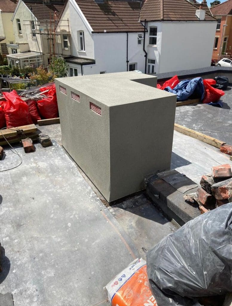 A rectangular, smooth-surfaced structure is on a rooftop surrounded by bricks, construction materials, and debris. Several red bags are stacked nearby, with residential houses visible in the background.