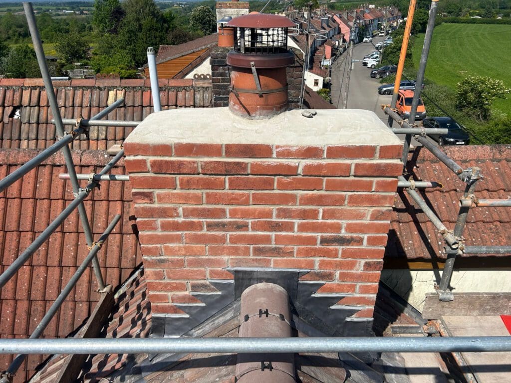 A newly constructed red brick chimney is surrounded by scaffolding on a rooftop. The chimney top has a metal cap. Below, the tiled roof is visible. In the background, a scenic view shows a line of houses, trees, and a grassy field.