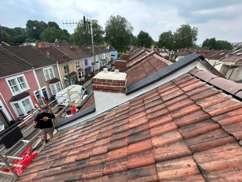 A view from a rooftop overlooking a row of terraced houses. The roofs are made of red and brown tiles, and chimneys are visible. A person stands on a ladder near the rooftops, with trees and cloudy sky in the background.