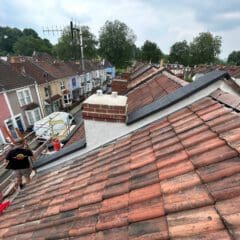 A view from a rooftop overlooking a row of terraced houses. The roofs are made of red and brown tiles, and chimneys are visible. A person stands on a ladder near the rooftops, with trees and cloudy sky in the background.