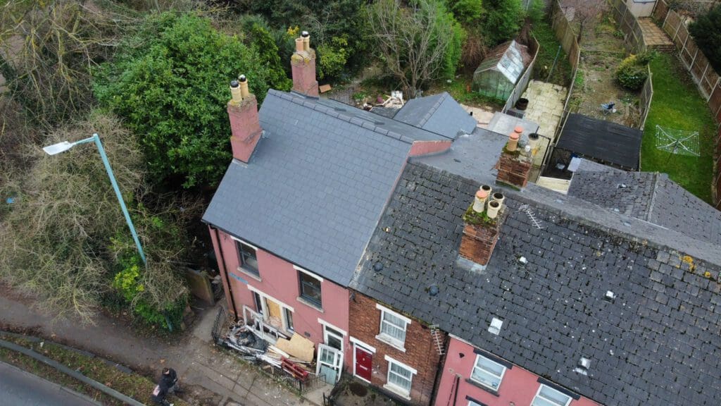 Aerial view of a row of pink and gray houses with dark roofs. The leftmost house shows signs of damage with debris near the entrance. Surrounding the houses are trees, a lamppost, a greenhouse, and a small backyard with scattered items.