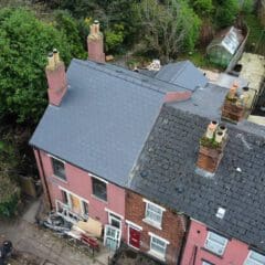 Aerial view of a row of pink and gray houses with dark roofs. The leftmost house shows signs of damage with debris near the entrance. Surrounding the houses are trees, a lamppost, a greenhouse, and a small backyard with scattered items.