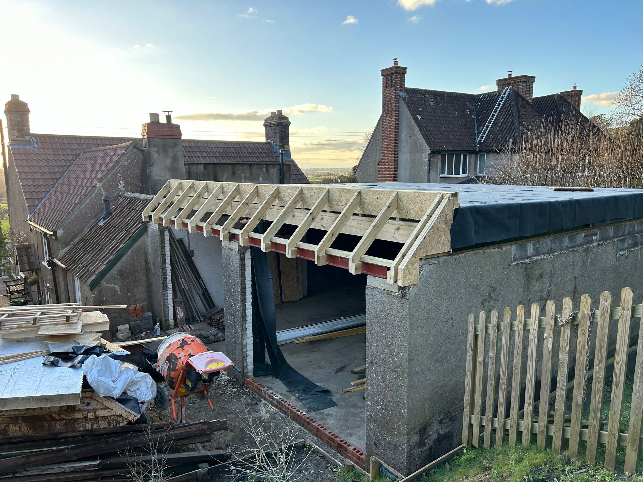 A construction site with a partially built garage attached to a house. The roof frame is exposed, and various building materials are scattered around, including wood and a cement mixer. A wooden fence is in the foreground, and houses are in the background.