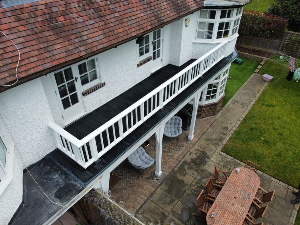 Aerial view of a two-story white house with a tiled roof. The upper floor features a balcony with a white railing. Below, there's a patio with a long dining table and chairs. A well-maintained garden with a lawn and pathway is visible.