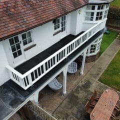 Aerial view of a two-story white house with a tiled roof. The upper floor features a balcony with a white railing. Below, there's a patio with a long dining table and chairs. A well-maintained garden with a lawn and pathway is visible.