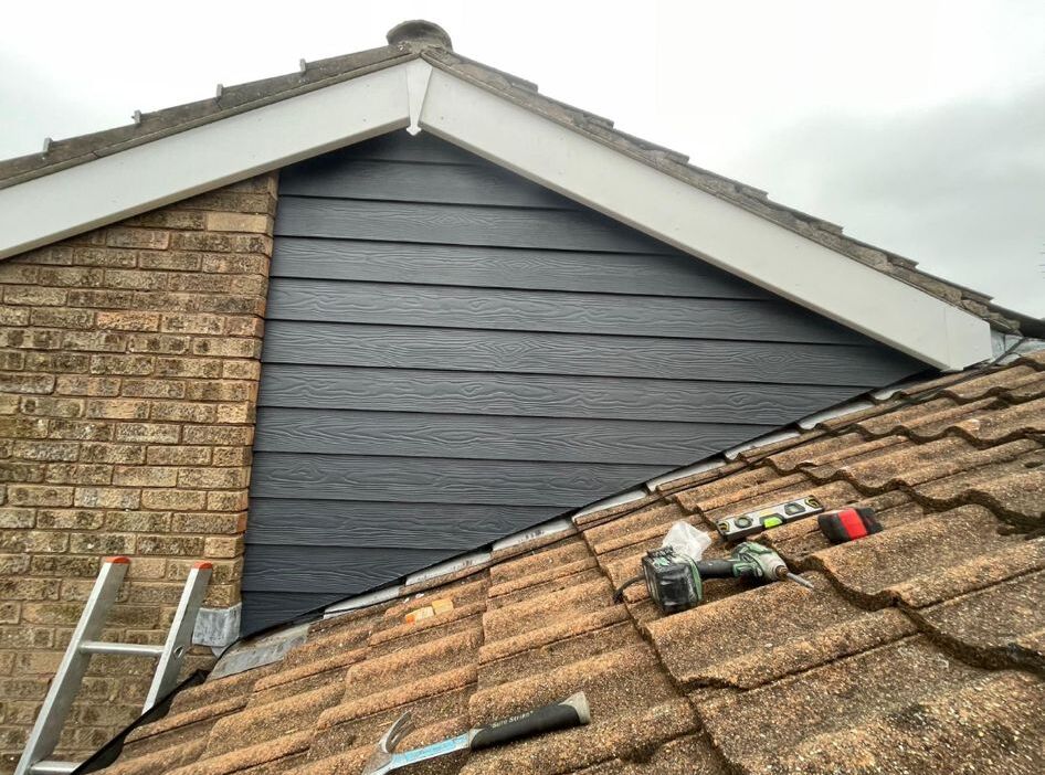 A roof with brown tiles partially covers a brick wall. The wall features a section of dark gray cladding. Tools including a hammer, drill, and adhesive are resting on the roof. A ladder leans against the building.